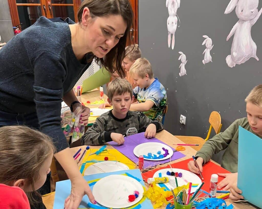 Zdjęcie przedstawia arteterapeutkę podczas pracy, która nachyla się nad stołem, wokół którego siedzą dzieci i wykonują pracę plastyczną
