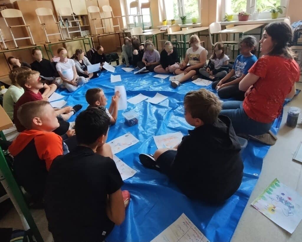 Jak wygląda sesja arteterapeutyczna. zdjęcie przedstawia dzieci siedzące w kręgu na podłodze, w sali szkolnej. Mają przed sobą rozłożone kartki i rysują. Wśród nich siedzi arteterapeutka. Wizualizacja odpowiedzi na pytanie: jak wygląda sesja arteterapeutyczna.