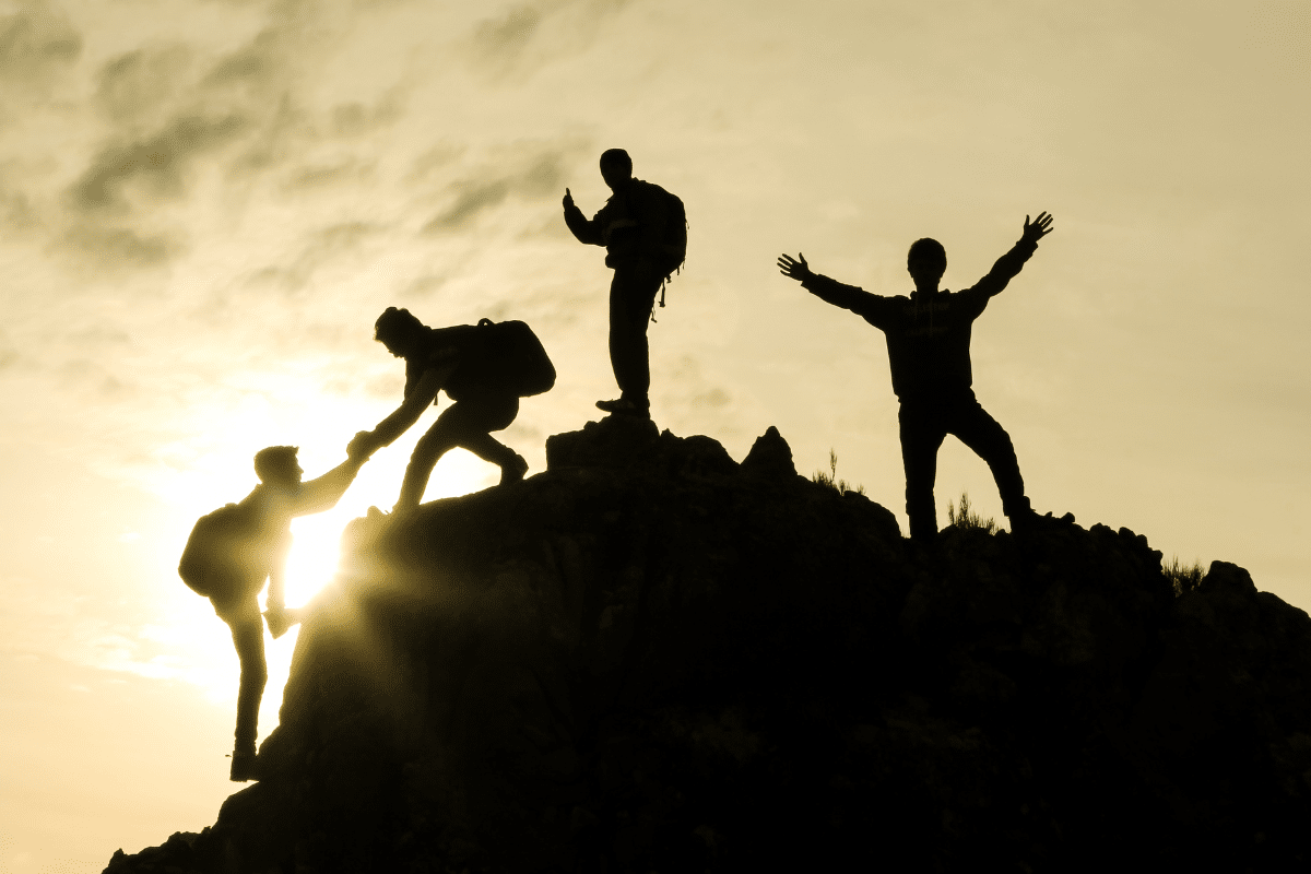 Zdjęcie przedstawiające pomagających sobie ludzi podczas wspinaczki na szczyt góry o wschodzie słońca.